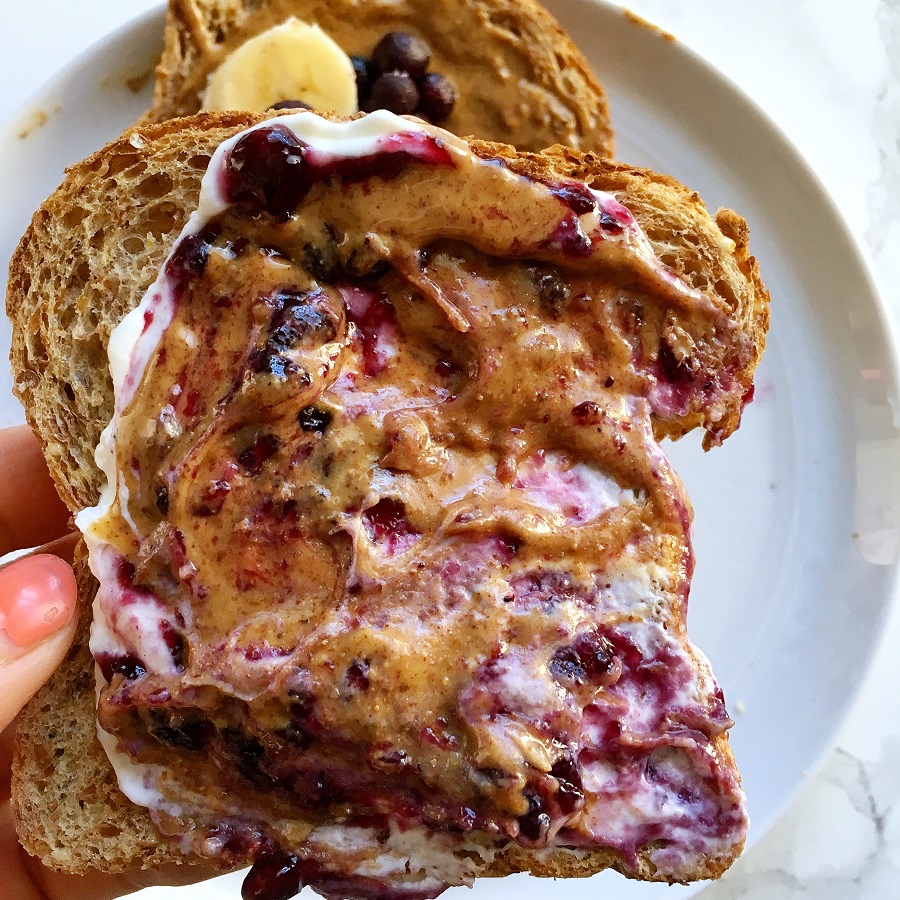 hand holding piece of bread topped with yogurt, peanut butter and jelly