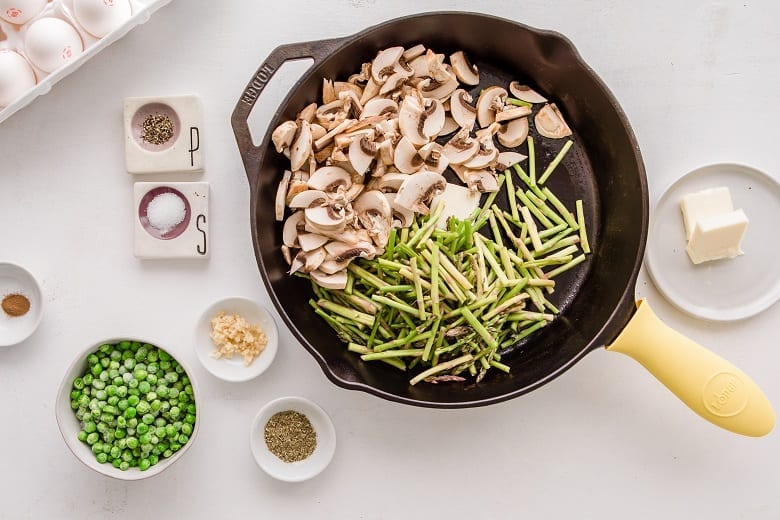 Skillet with asparagus and mushrooms on white table with bowl of peas and spices