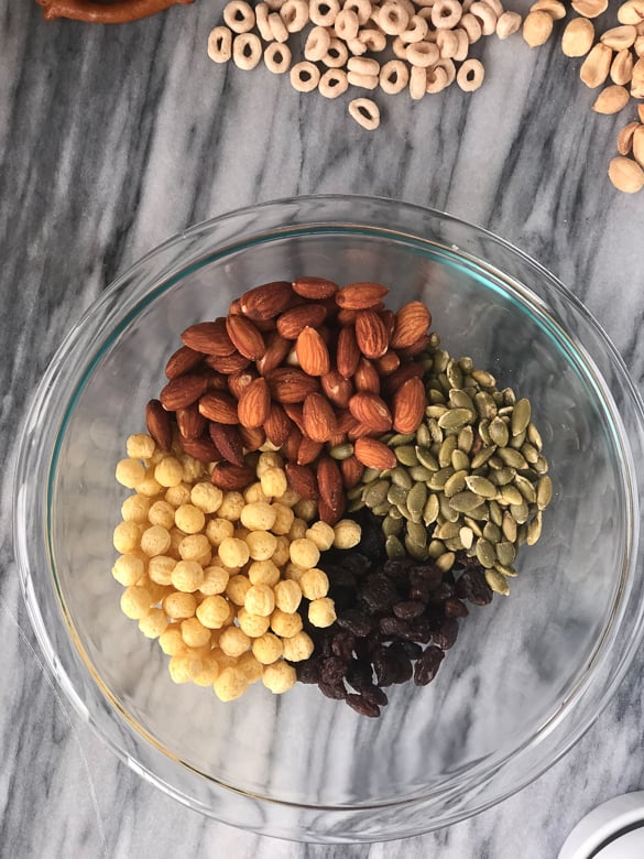 pumpkin seeds, almonds, kix and raisins in a bowl for homemade trail mix in a clear mixing bowl