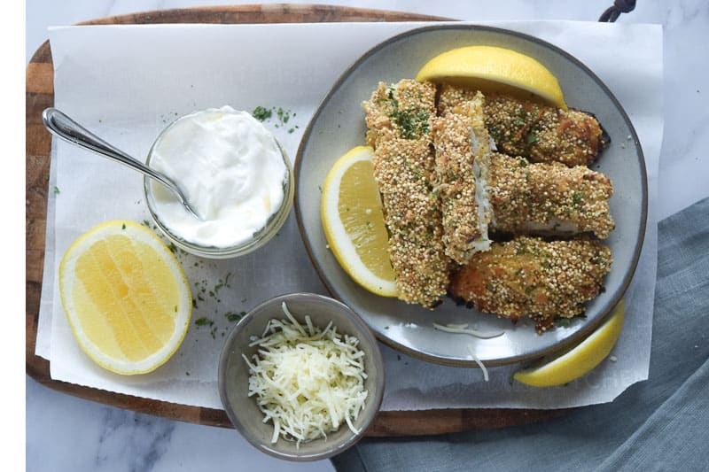 Healthy gluten free fish sticks with quinoa breading on small plate with lemons and side of yogurt dipping sauce 