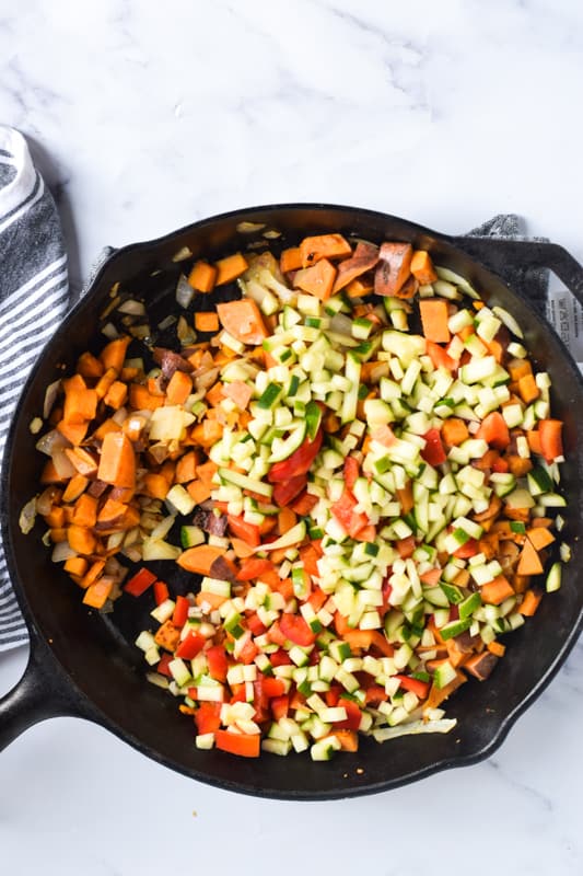 Sauteed onions, zucchini, tomatoes and sweet potatoes in cast iron skillet for vegan breakfast hash. 
