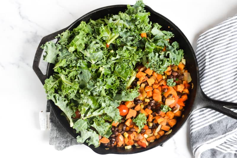 Veggie hash in skillet with raw kale on white countertop with striped napkin
