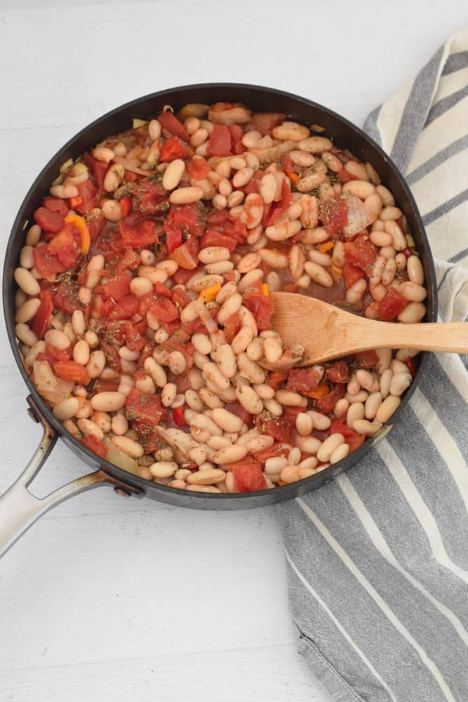 skillet with white beans and tomatoes with wooden spoon over blue striped napkin