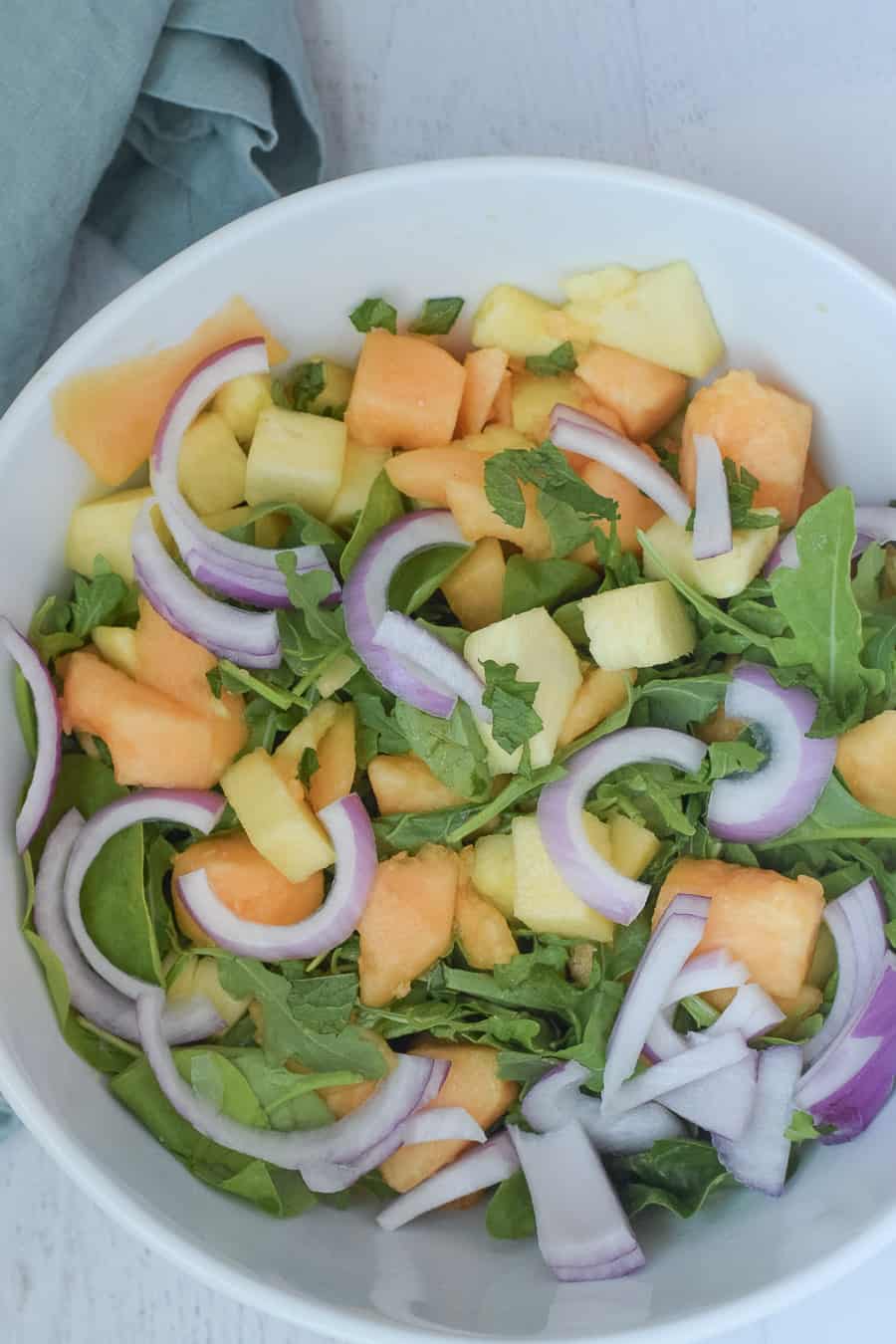 Cantaloupe salad with pineapple and red onion over bed of arugula greens in white salad bowl